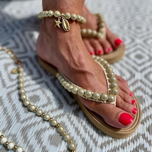 Könnte beinhalten: Ein Paar beige Flip-Flops mit Perlenbesatz. Die Flip-Flops haben ein goldenes Fußkettchen mit einem Seestern- und Muschel-Anhänger.