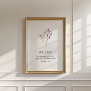 May include: A framed wall art print featuring a watercolor illustration of small purple flowers. Below the flowers, the text reads in Arabic script, followed by the English text: SO REMEMBER ME, I WILL REMEMBER YOU. QURAN 2:152.