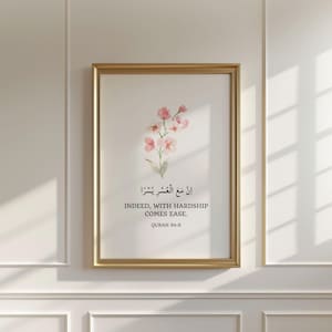 May include: A framed wall art print featuring a watercolor illustration of pink flowers above Arabic calligraphy and the English text: "INDEED, WITH HARDSHIP COMES EASE. QURAN 94:6". The print is enclosed in a thin gold frame with a white background.