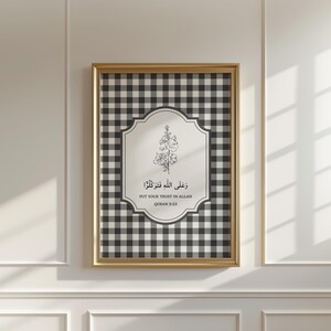 Puede incluir: Obra de arte enmarcada con un marco dorado. Presenta un fondo a cuadros vichy en blanco y negro. En el centro, un panel blanco con ilustraciones florales y texto árabe: "Confía en Allah, Corán 5:23".