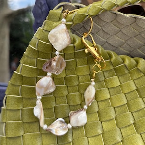 Puede incluir: Un bolso tejido verde con una cadena dorada y un dije hecho de piezas de concha blanca.
