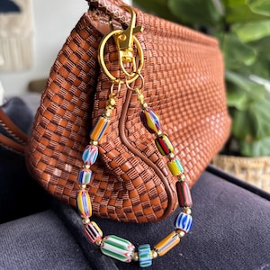 May include: A brown woven leather handbag with a gold-colored clasp and a beaded charm. The charm features colorful, cylindrical beads in various colors, including blue, green, yellow, and red. The handbag's texture is visible.