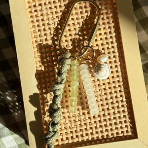 May include: A gold-colored keychain with a woven olive green lanyard. The keychain features a series of light green and white beads, along with a small seashell charm. The background is a light brown woven pattern.