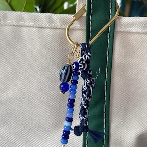 May include: A gold-toned key chain with a blue and white beaded charm and a blue and white fabric charm. The key chain is attached to a white canvas tote bag with a green strap.