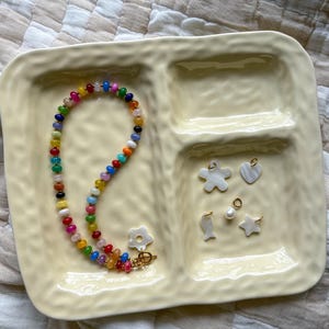 May include: A yellow divided tray holds a colorful beaded necklace and several small charms. The necklace features a variety of colorful beads. Charms include a flower, heart, fish, star, and a pearl. The tray is on a patterned surface.