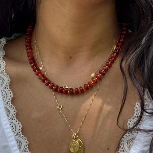 May include: A layered gold necklace with a red carnelian bead strand and a pendant featuring two gold medallions.