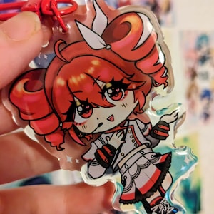 May include: A clear acrylic charm depicting a cartoon character with red hair and eyes, dressed in a white and red outfit. The charm is attached to a red clasp. The charm has a holographic effect.