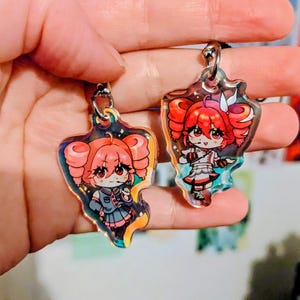 May include: Two anime-style keyrings featuring a character with red hair and eyes. The keyrings have a clear, iridescent background and a silver keyring. The character is wearing a blue and grey outfit on one keyring and a white and red outfit on the other.