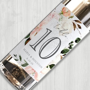 May include: A white wine bottle with a floral design label. The label features a large number 10, the text "table", and the names "Maria & Houston" with the date "6.10.20".