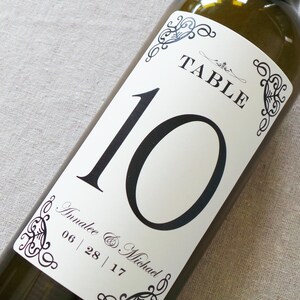 May include: A white table number card with black text and a decorative border. The card is attached to a brown glass wine bottle. The card reads "Table 10" and "Annalee & Michael 06 | 28 | 17".