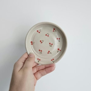 Puede incluir: Un plato pequeño y redondo de cerámica blanquecina decorado con cerezas rojas pintadas a mano. Las cerezas están dispersas por la superficie del plato. El plato está sostenido en una mano.