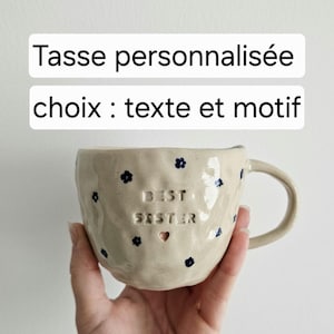 Può includere: Tazza in ceramica beige con superficie strutturata, decorata con piccoli disegni floreali blu. La tazza presenta le parole "BEST SISTER" e un simbolo a forma di cuore. Il testo "Tasse personnalisée choix : texte et motif" è sopra.