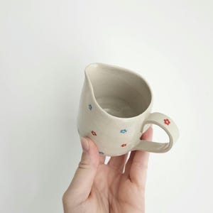 Puede incluir: Una jarra de cerámica beige con un pico y asa curvados. La jarra está decorada con pequeños diseños de flores rojas y azules. El asa también tiene un diseño de flor roja. La jarra está sostenida por una mano.