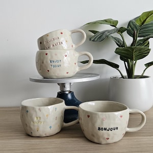 Peut inclure: Ensemble de tasses en céramique faites à la main, de couleur crème. Les tasses présentent des surfaces texturées, des motifs de cœurs et de fleurs, et des phrases comme "Enjoy Today" et "Bonjour". Les tasses sont exposées avec une plante en pot en arrière-plan.