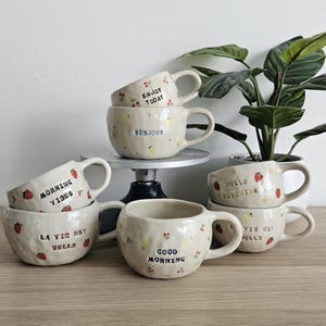Peut inclure: Plusieurs tasses en céramique faites à la main, de tailles et de motifs variés. Les tasses blanc cassé ont une surface texturée et présentent des détails peints à la main, comme des fraises, des cerises et des phrases.