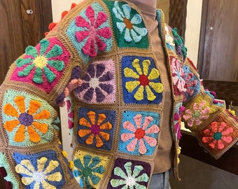 Puede incluir: Chaqueta de ganchillo hecha a mano, colorida, con un diseño de patchwork. Cada panel cuadrado muestra un motivo floral vibrante en tonos rosa, azul, amarillo y naranja, sobre un fondo marrón. La chaqueta tiene mangas largas y cuello.