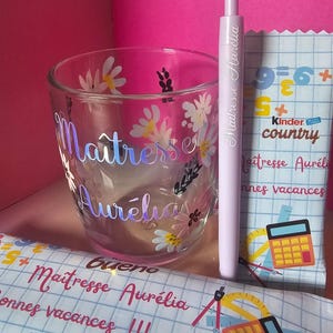 Op de afbeelding: Een helder glas met bloemmotieven en de naam "Maîtresse Aurélia". Een lichtpaarse pen en een Kinder Country chocoladereep zijn ook zichtbaar. De reep heeft tekst en een rekenmachine-afbeelding.