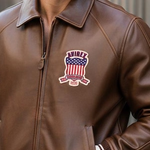 May include: A brown leather jacket with a zipper closure, side pockets, and a white and black striped trim on the cuffs and waist. The jacket features a patch on the chest with the text "AVIREX" and an American flag design. The person in the image is wearing black sunglasses.