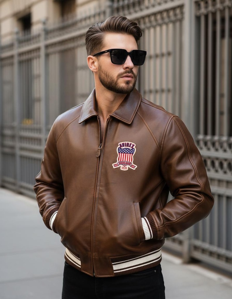 May include: A brown leather jacket with a zipper closure, side pockets, and a white and black striped trim on the cuffs and waist. The jacket features a patch on the chest with the text "AVIREX" and an American flag design. The person in the image is wearing black sunglasses.