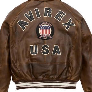 Puede incluir: Chaqueta de cuero marr&oacute;n con la palabra "AVIREX" en letras blancas en la espalda. Debajo, un parche con "ALL AMERICANS" y "ESTABLISHED 1975" alrededor de un dise&ntilde;o de la bandera estadounidense. La palabra "USA" est&aacute; debajo. La chaqueta tiene un dobladillo acanalado.