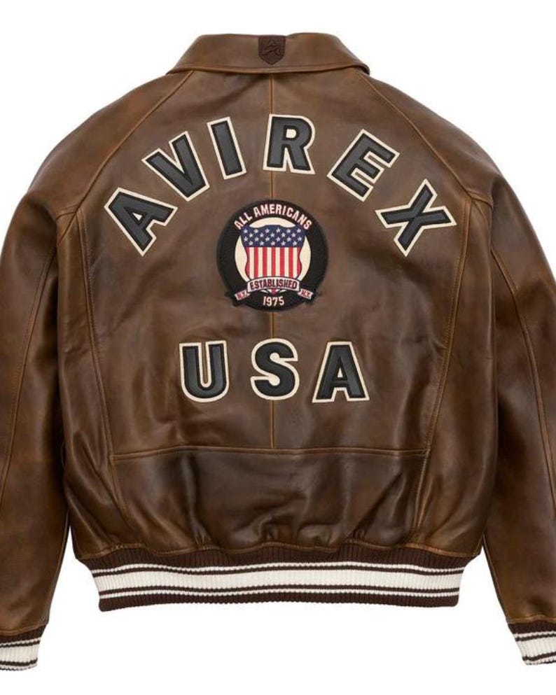 Puede incluir: Chaqueta de cuero marr&oacute;n con la palabra "AVIREX" en letras blancas en la espalda. Debajo, un parche con "ALL AMERICANS" y "ESTABLISHED 1975" alrededor de un dise&ntilde;o de la bandera estadounidense. La palabra "USA" est&aacute; debajo. La chaqueta tiene un dobladillo acanalado.