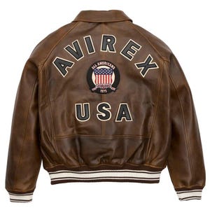 Puede incluir: Chaqueta bomber de cuero marr&oacute;n con la palabra "AVIREX" en la espalda, junto con un parche que dice "ALL AMERICANS ESTABLISHED 1975" y "USA". La chaqueta tiene cuello y pu&ntilde;os acanalados con rayas blancas.