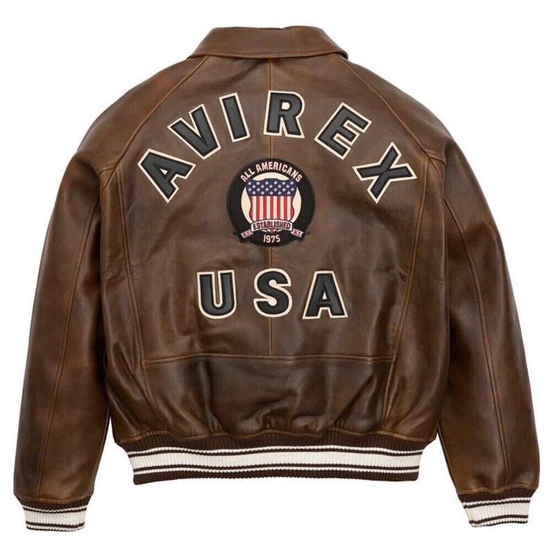 Puede incluir: Chaqueta bomber de cuero marr&oacute;n con la palabra "AVIREX" en la espalda, junto con un parche que dice "ALL AMERICANS ESTABLISHED 1975" y "USA". La chaqueta tiene cuello y pu&ntilde;os acanalados con rayas blancas.