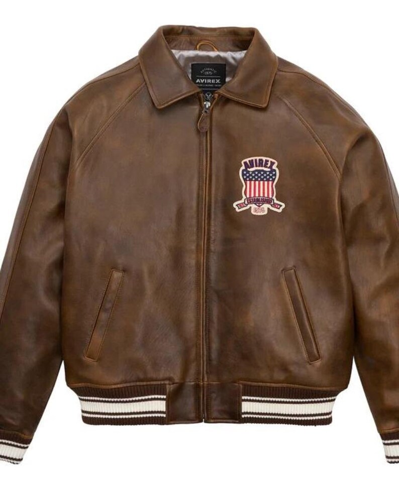 Puede incluir: Chaqueta de cuero marr&oacute;n de estilo bomber. Tiene cuello, cierre de cremallera y dos bolsillos delanteros. Un parche bordado con un escudo est&aacute; en el pecho. Los pu&ntilde;os y el dobladillo tienen un patr&oacute;n de rayas.