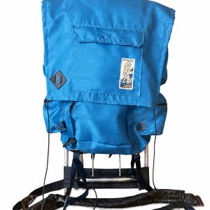 Puede incluir: Una mochila de marco externo azul brillante con un compartimento principal grande con cierre de solapa, dos bolsillos laterales y un parche rectangular con el texto CONQUEST. La mochila está unida a un marco de metal plateado con correas negras.