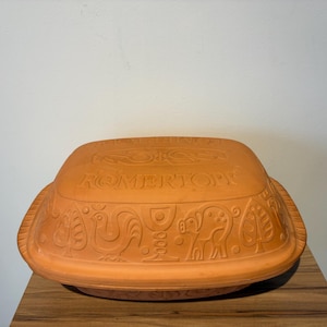 May include: A terracotta-colored Römertopf clay baker with a rectangular shape and a lid. The lid and sides feature embossed decorative patterns and the word "Römertopf". The baker sits on a wooden surface.