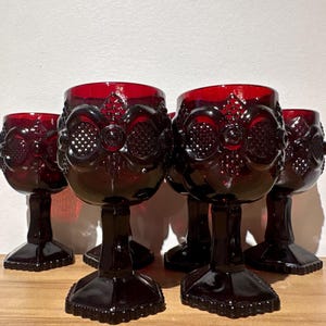 Peut inclure: Un ensemble de cinq verres à pied en verre rouge profond présentant un motif en relief composé de cercles et de losanges. Chaque verre possède un pied épais à facettes et une base carrée, avec un design en verre pressé de couleur rubis.