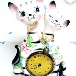 May include: A vintage ceramic cat clock featuring two cat figures. The white cats have black stripes, pink cheeks, and blue eyes. The clock face is yellow with black numbers and hands, and is decorated with floral accents.