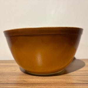 May include: A round mixing bowl with a smooth, glossy finish in a solid, deep amber-brown colour. The bowl features a wide, slightly flared rim and a curved body that tapers down to a smaller, circular base, presenting a simple, functional design.