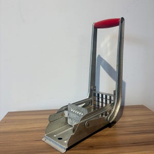 May include: A vintage, silver-toned potato cutter with a red handle. The device features a grid-like blade for slicing potatoes and a hinged design for easy use. The cutter is made of metal and sits on a wooden surface.