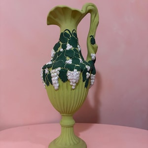 May include: A light green ceramic vase with a fluted body and a pedestal base. The vase is adorned with dark green leaves and white grape clusters. The handle curves upwards. The vase is a decorative item.