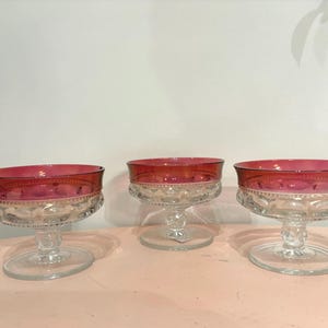 Peut inclure: Trois coupes à dessert vintage en verre, avec des bords rouge rubis et des bases transparentes ornées. Les coupes sont disposées sur une surface rose clair, mettant en valeur leur design élégant. Idéales pour les desserts.