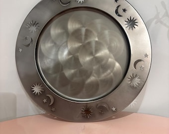 May include: A circular metal tray with a brushed finish featuring a central area with overlapping circular patterns. The wide rim is decorated with cut-out celestial motifs including suns, crescent moons, and small stars arranged in a repeating pattern.