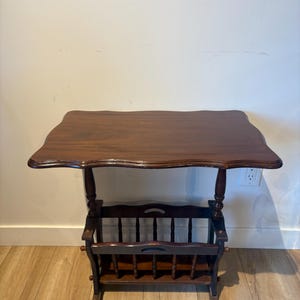 May include: A dark brown wooden side table featuring a rectangular top with scalloped edges. The table includes a lower storage rack constructed with vertical spindles and a handle cutout on each side, supported by four turned wooden legs.
