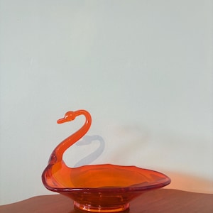 May include: An orange glass swan-shaped dish. The swan has a long, curved neck and a wide, shallow bowl. The dish is made of transparent glass and sits on a round base. The dish is likely vintage and is a decorative item.