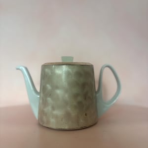 May include: A ceramic teapot with a light grey body and light blue handle, spout, and lid. The teapot has a textured surface and a modern design. The background is a solid light pink.