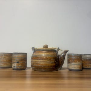 Peut inclure: Service à thé en céramique avec une théière et quatre tasses assorties. L'ensemble présente un motif rayé marron et beige. La théière a une anse et un couvercle en bois. Les tasses sont cylindriques.