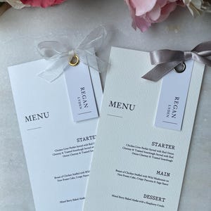 May include: Two white wedding menu cards with menu details and names, tied with ribbons and tags. The menus feature the names "Regan Lyden" and "Lucie + Paul" with the date "07.04.2025".