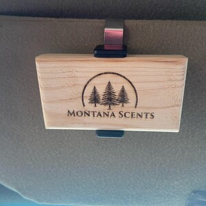May include: A rectangular wooden car air freshener with the text "MONTANA SCENTS" and a circular graphic of three pine trees. The air freshener is attached to a metal clip and a black plastic piece.