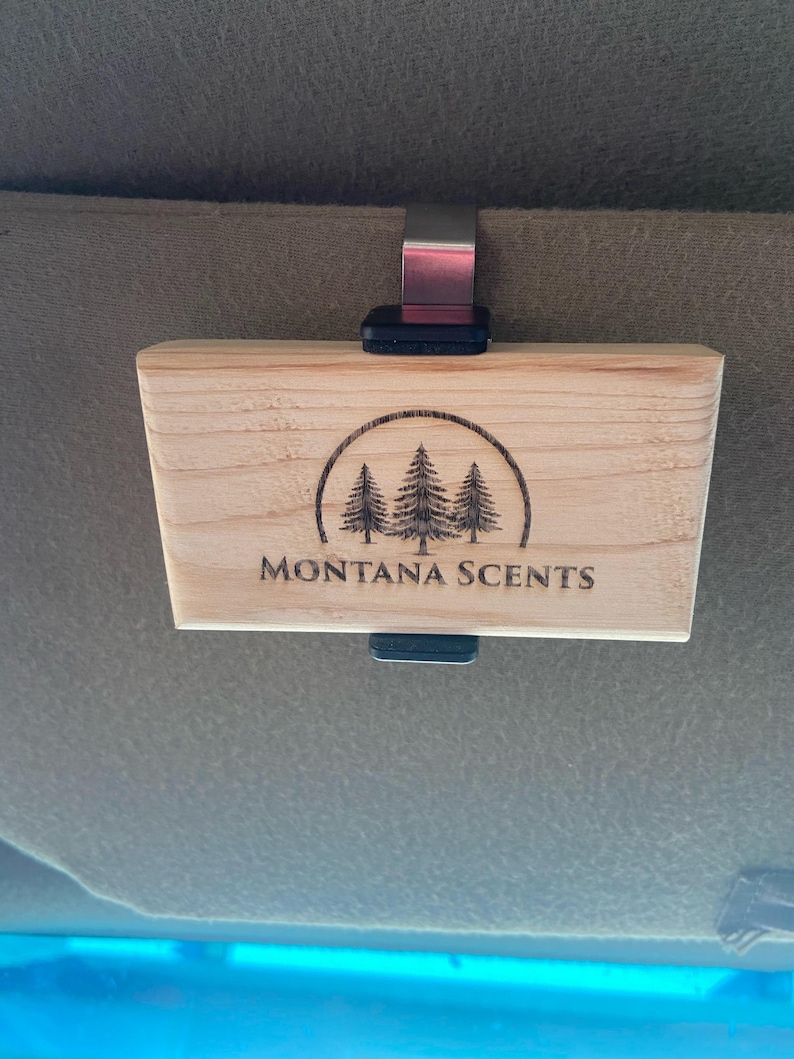 May include: A rectangular wooden car air freshener with the text "MONTANA SCENTS" and a circular graphic of three pine trees. The air freshener is attached to a metal clip and a black plastic piece.