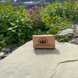 May include: A small, rectangular wooden box with the text "MONTANA SCENTS" and a tree logo is displayed on a rock. The background features a mountain range, green foliage, and colorful wildflowers under a cloudy sky.