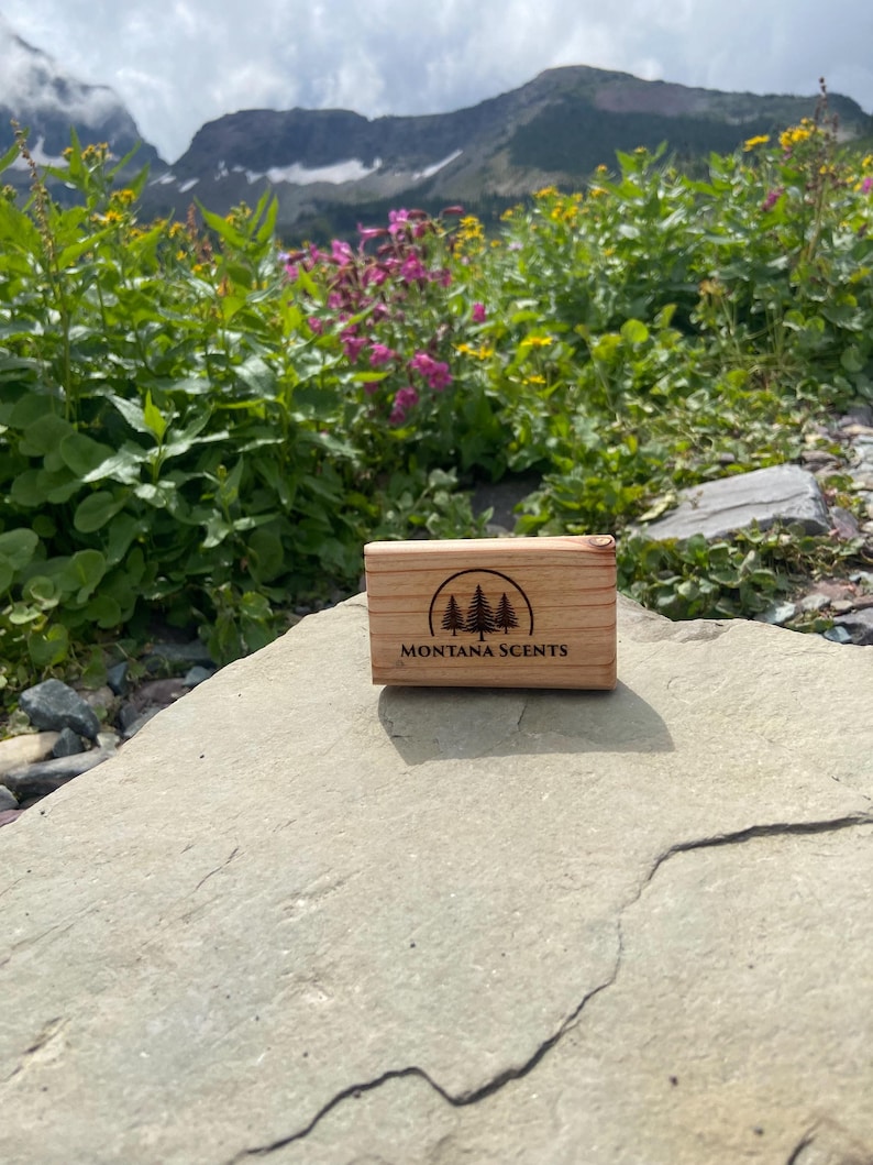 May include: A small, rectangular wooden box with the text "MONTANA SCENTS" and a tree logo is displayed on a rock. The background features a mountain range, green foliage, and colorful wildflowers under a cloudy sky.