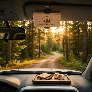 May include: A wooden car air freshener with the text "MONTANA SCENTS" and a tree logo is attached to the car's visor. A wooden tray on the dashboard holds s'mores. The background shows a forest road.