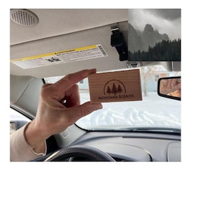 May include: A rectangular wooden air freshener with the text "MONTANA SCENTS" and a tree logo is held in front of a car's interior. The car's visor and a mountain scene are visible in the background. The air freshener is made of light-colored wood.