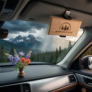 May include: A wooden car air freshener with the text "MONTANA SCENTS" and a tree logo, hanging from a car visor. A small vase of colorful wildflowers sits on the dashboard. The background shows a mountain range and forest.