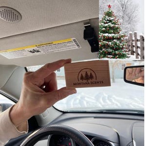 May include: A rectangular wooden air freshener with the text "MONTANA SCENTS" and a tree logo is held in a car. The car's interior is visible, along with a Christmas tree in the background. The air freshener is light brown.